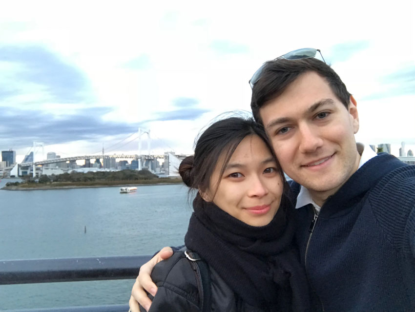 A girl and boy, smiling. They are both wearing dark clothes. The girl has a big black scarf around her neck. The boy has sunglasses on top of his head and his arm around the girl. In the background is a view of a skyline on the water, and a bridge.