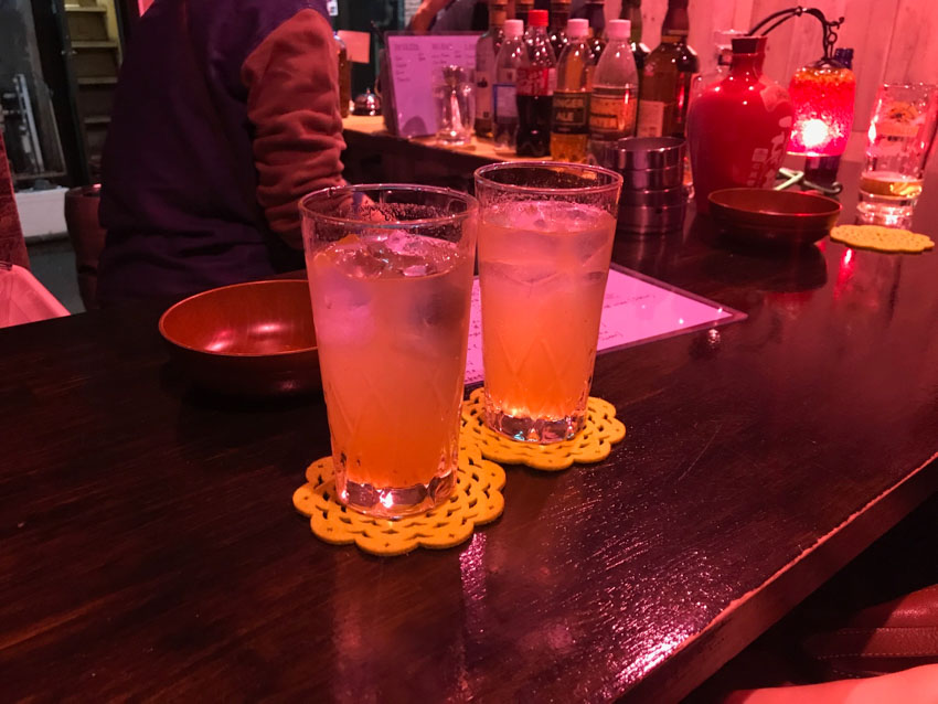 Two glasses of umeshu plum wine and soda on yellow flower-shaped coasters, sitting on a wooden tabletop in a dimly lit bar