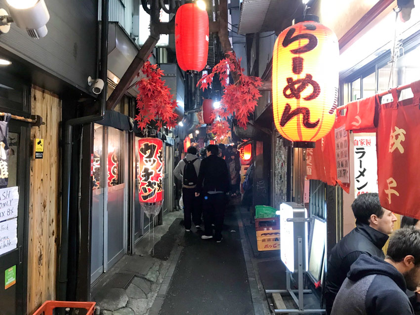 A narrow alleyway with people walking several steps ahead. Red lanterns with Japanese hiragana printed on them hang from some restaurant fronts, people are seen on the right, seated in the restaurants but close to the outside of the restaurant