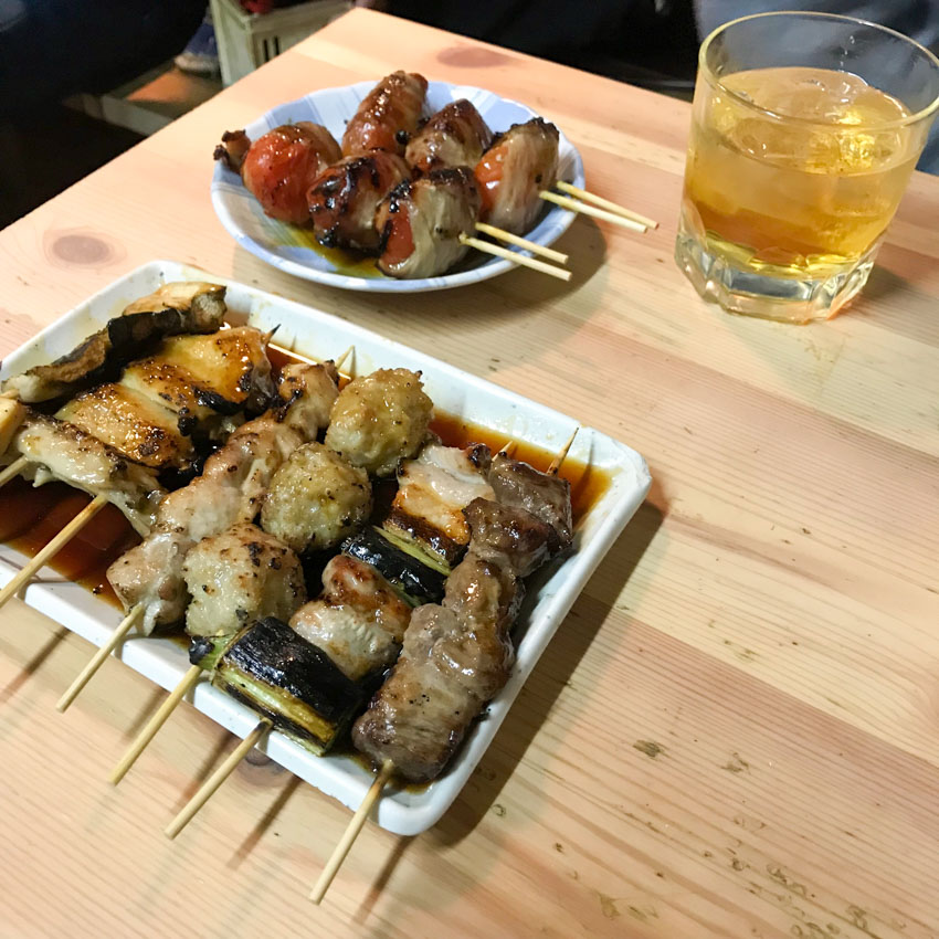 A round dish and rectangular dish on a light wooden table, filled with meat and vegetable skewers, doused in sauce. A short tumbler of umeshu plum wine sits on the table