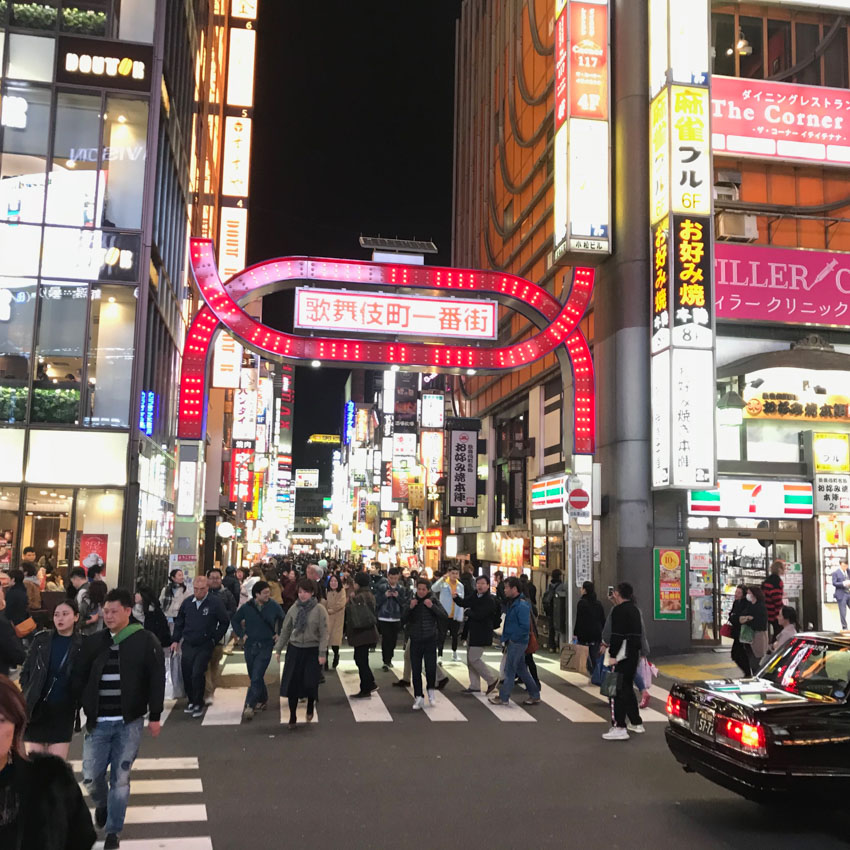 A busy entrance to Kabukichō, off the main road, with a giant pink neon arch as signage. The entrance sits between two high-rise buildings which also have neon signs. Many people cross the crossings that run across the entrance
