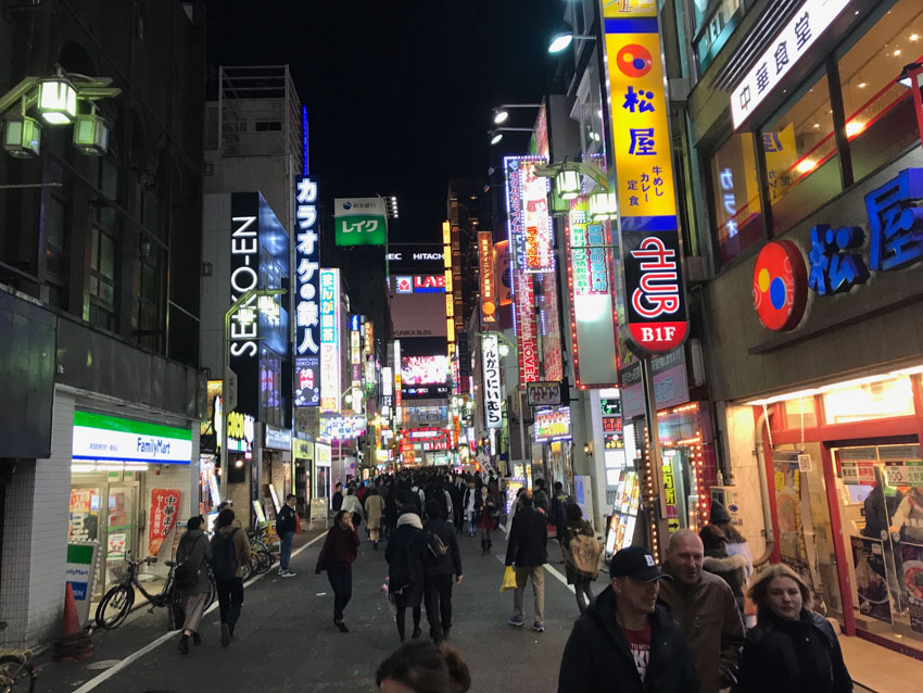 A street at night time, fairly busy with people, with stores and restaurants highlighted by big neon signs