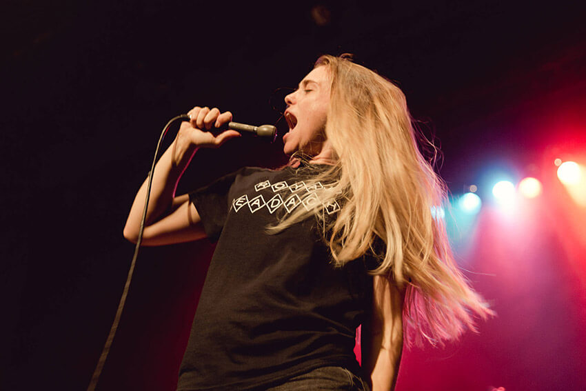 A woman with blonde hair singing loudly into a hand-held microphone on stage