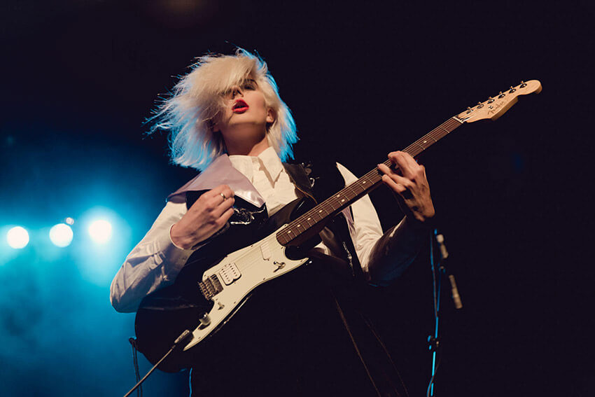 A low shot of a woman with pale coloured hair and red lips, playing an electric guitar with blue lights in the background