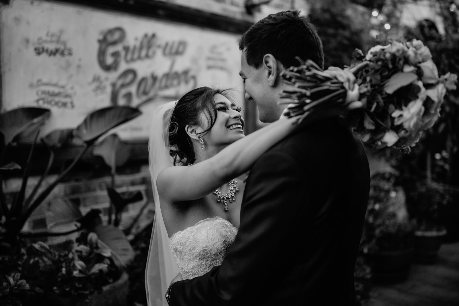 A black and white image of a man and woman embracing on their wedding day. The woman has dark hair in an up-do and a veil hanging from her bun, she has her arms around the man’s neck as she holds a bouquet of flowers, both the man and woman are smiling. In the background is an old-style billboard with cursive text on it, and some plants.