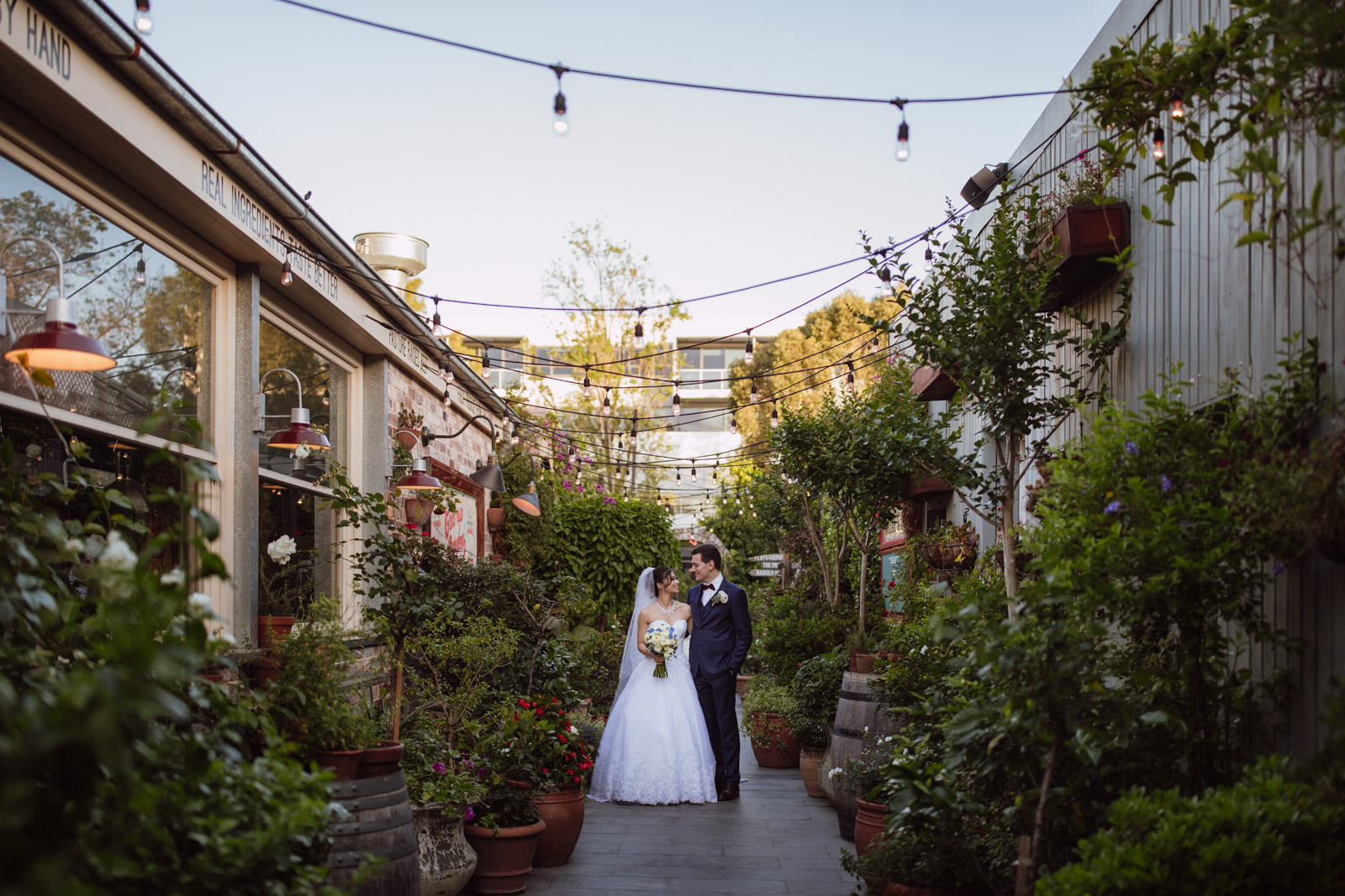 A man and woman on their wedding day in a narrow alley lined with a lot of pot plants. Light bulbs hang from power lines strung across the two buildings on either side. The man and woman are looking at each other and smiling. The woman is wearing a white strapless ballgown and a white veil, and is holding a bouquet of ivory and blue flowers. The man is wearing a navy blue suit