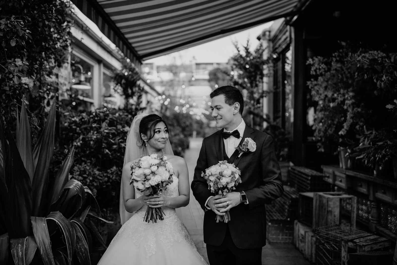 A black and white photo of a man and woman on their wedding day, both holding bouquets of roses. In the background are some plants and wooden crates.