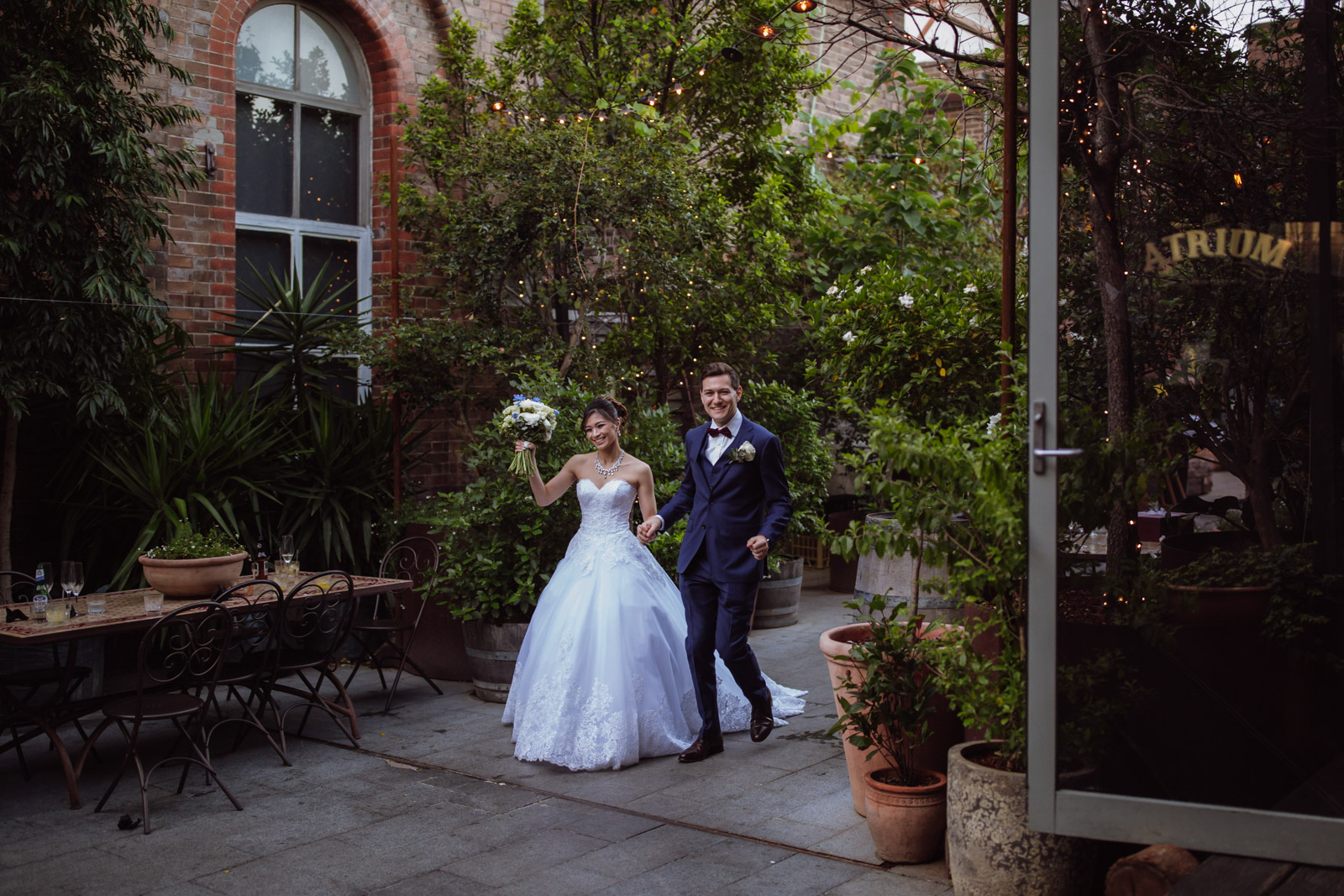 A man and woman walking holding hands, smiling. The woman is wearing a white wedding ballgown and the man is wearing a navy blue suit. In the background is a wooden table with some ornate metal chairs. The table has some wine glasses on them. In the background is a building and a lot of pot plants, large and small. In the foreground is an open glass door reading “The Atrium”.