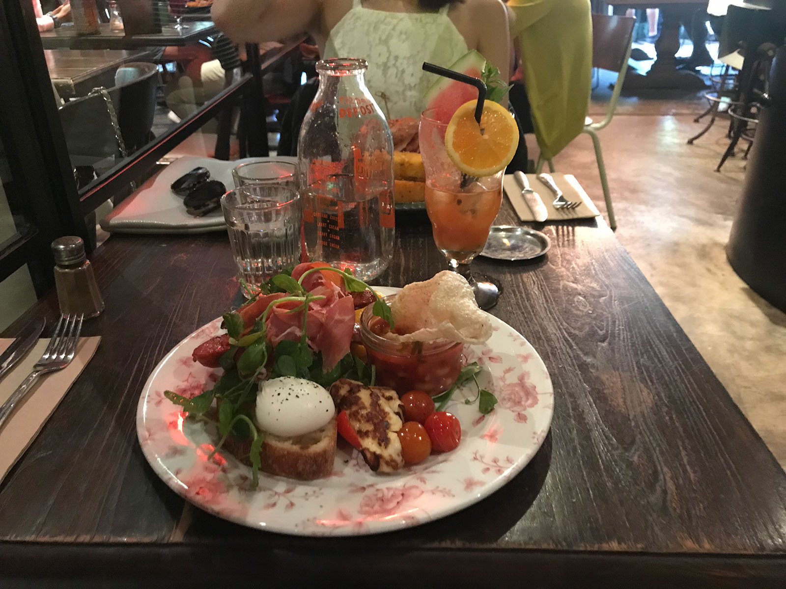 Toast topped with a poached egg, greens, prosciutto and chorizo, with small tomatoes, capsicum and halloumi on the side, served on a white plate with a floral design.
