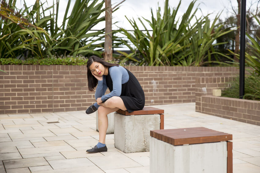 The same woman from other photos on this webpage, sitting on a concrete cube with a wooden top. She has the ankle of leg propped up on her opposite thigh and is leaning forward using her arm on her lap as balance. Other concrete cubes surround the one she is sitting on.