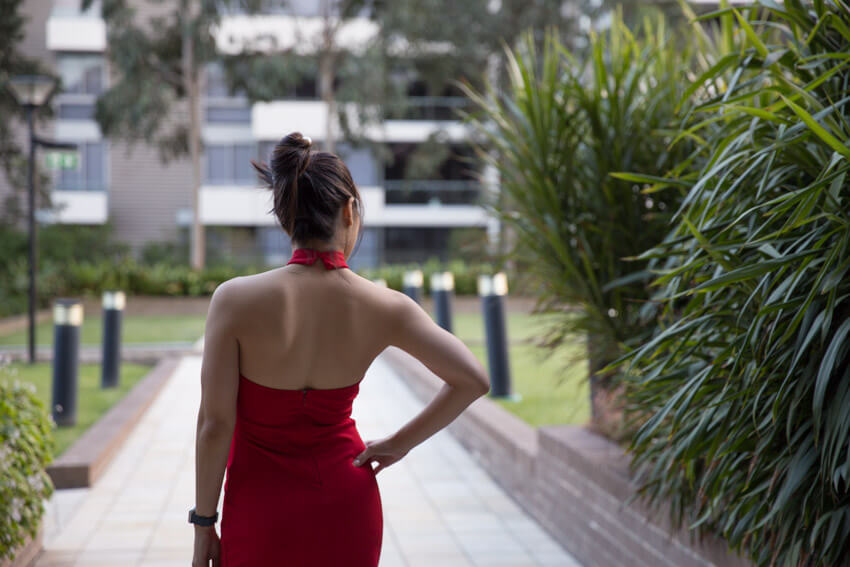 The same woman in the other photos on this webpage, facing away from the camera. She is wearing a red halter dress that shows her upper back. Her right hand is on her hip and her hair is up in a bun.