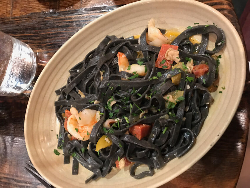 A beige oval bowl filled with black squid ink spaghetti, some prawns, and a dash of herbs
