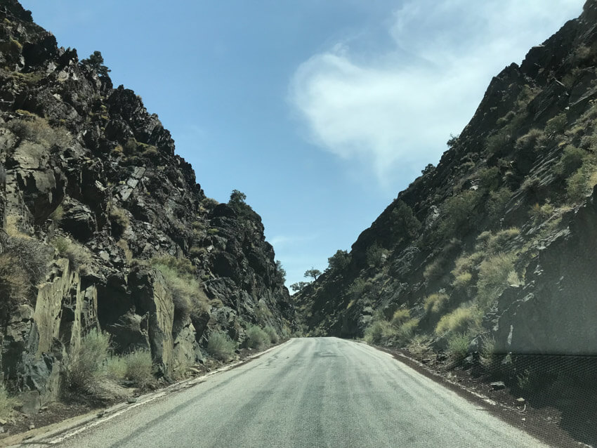 An unmarked road leading into the distance, lined at the sides with high formations of dark, almost black rock. There are weeds in the rocks.