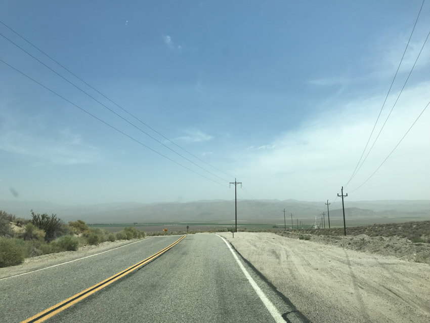 A stretch of road in a desert-like area, curving slightly to the left. There are power poles at the side of the road, with power lines connecting them to other power poles.