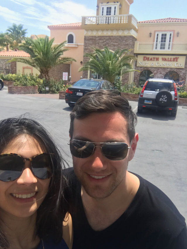 A man and woman wearing sunglasses, smiling. They are in a car park in front of a building that reads “Death Valley Nut & Candy Co.”