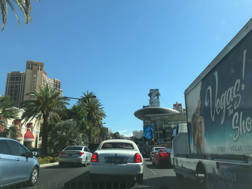 A multi-lane road with some traffic, as seen from the view of a car. There is a vehicle to the right with a billboard on it that reads “Vegas!”. In the distance are palm trees down the left of the road, and some tall hotels.