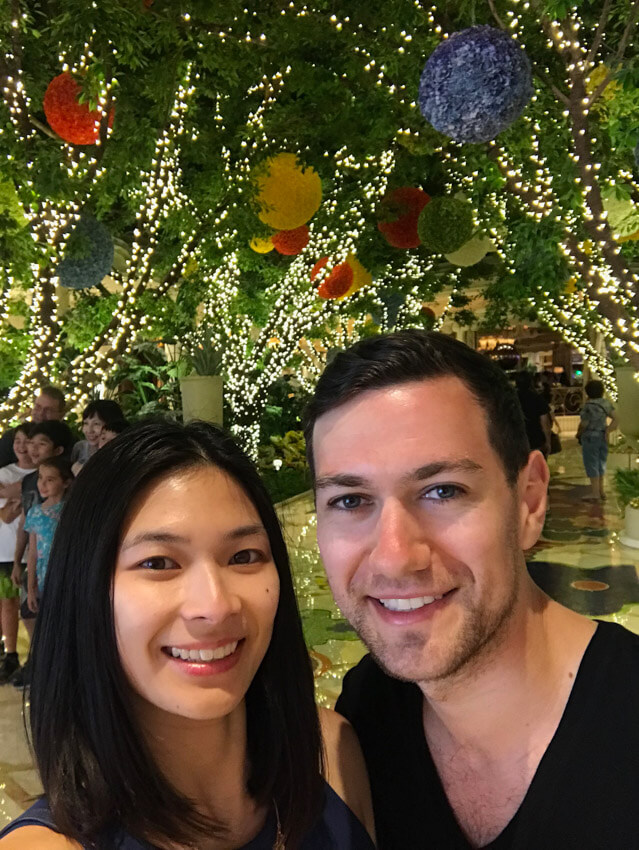 A man and woman smiling, with trees in the background that are decorated with fairy lights