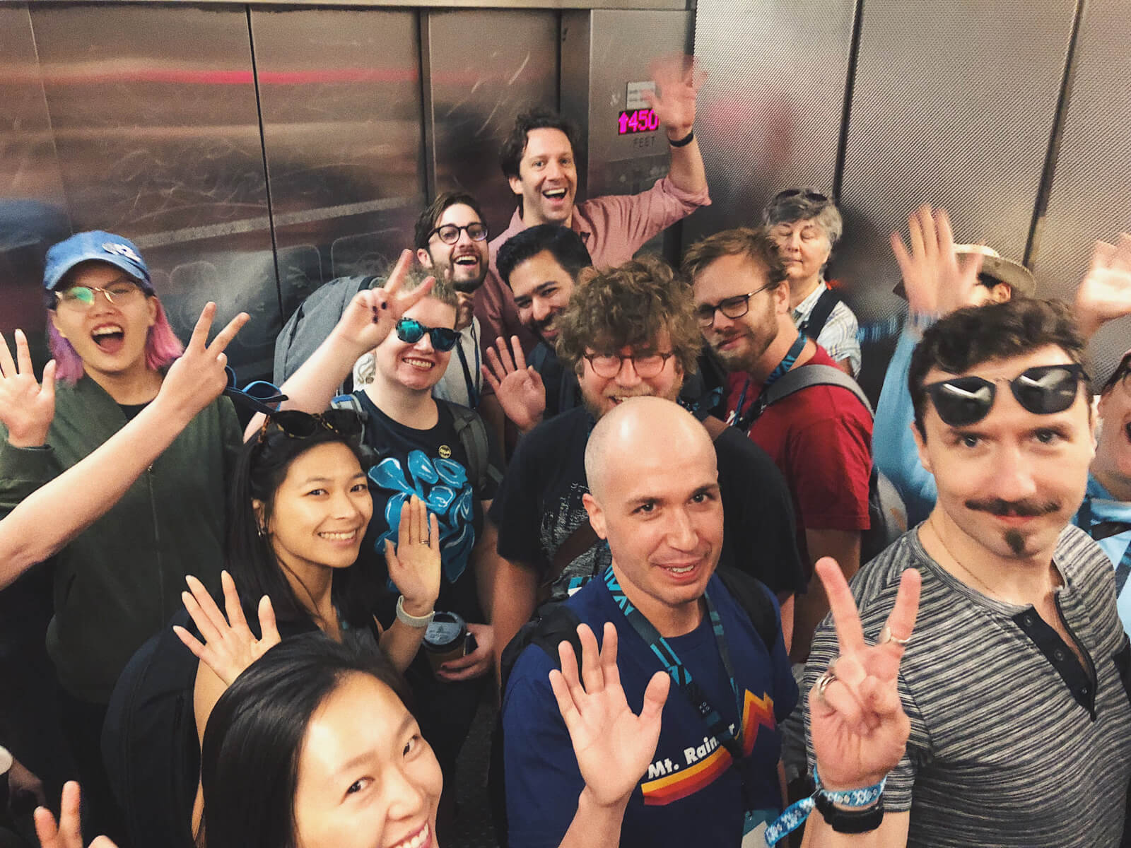 A handful of people in an elevator, all of them smiling/waving.