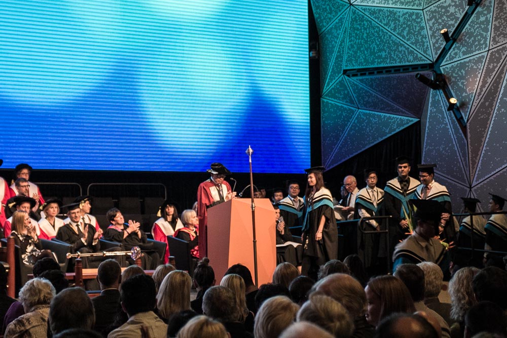 A university graduation ceremony on a stage, viewed from the crowd. Many people in blue gowns are lined up with one woman walking to the podium