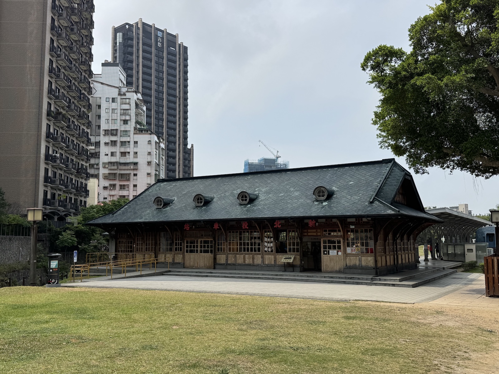 The restored wooden Xinbeitou Historic Station stands on a grassy plaza, its dark tiled roof and Japanese-era design framed by modern apartment towers.