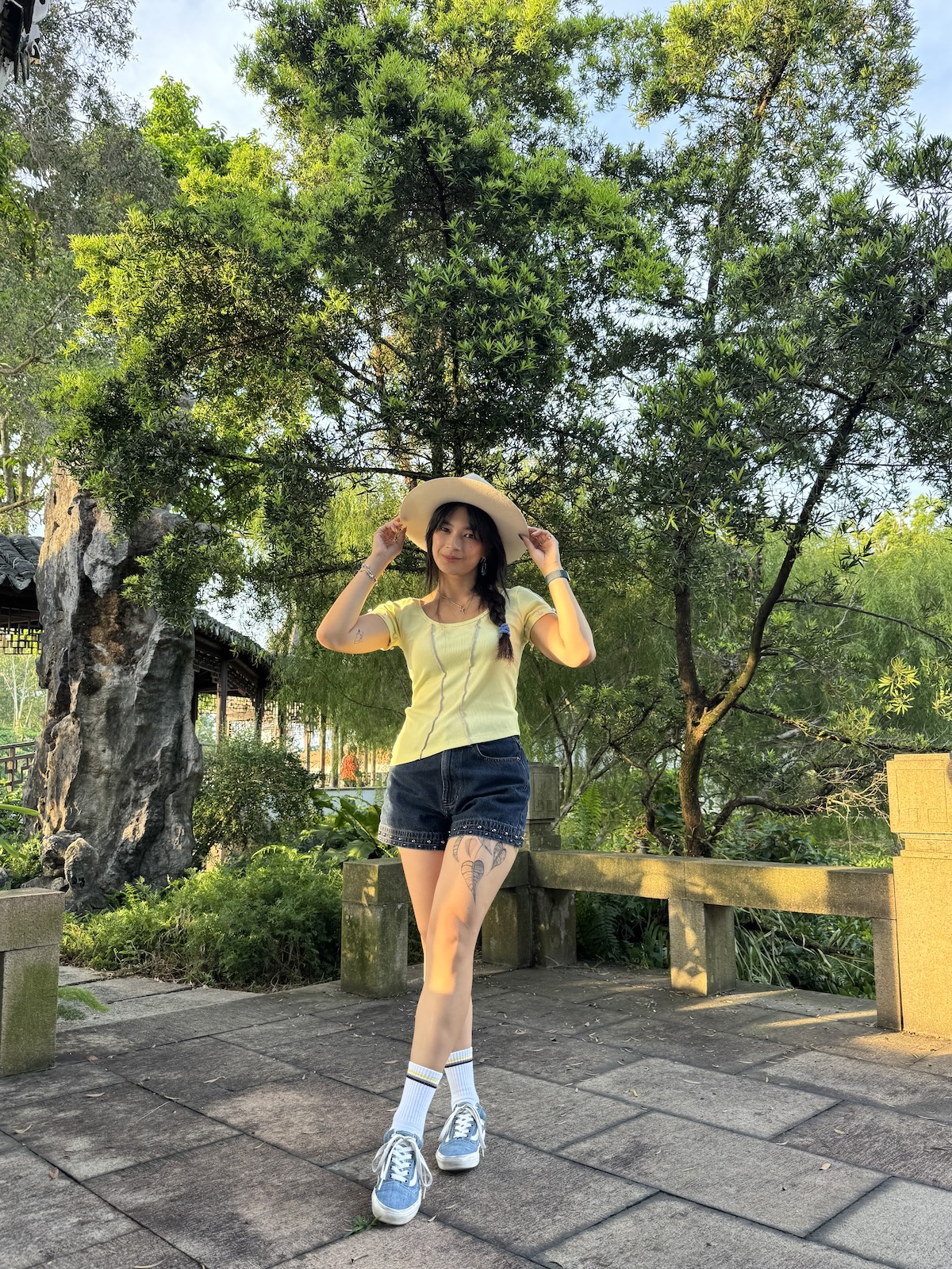 Me wearing a white sunhat, yellow short-sleeved top with light purple stitching detail, denim shorts with studs, white non-binary (yellow, black, purple) socks, and blue sneakers posing in the Japanese garden. I am holding the brim of my hat lightly with both hands and have one ankle crossed in front of the other.