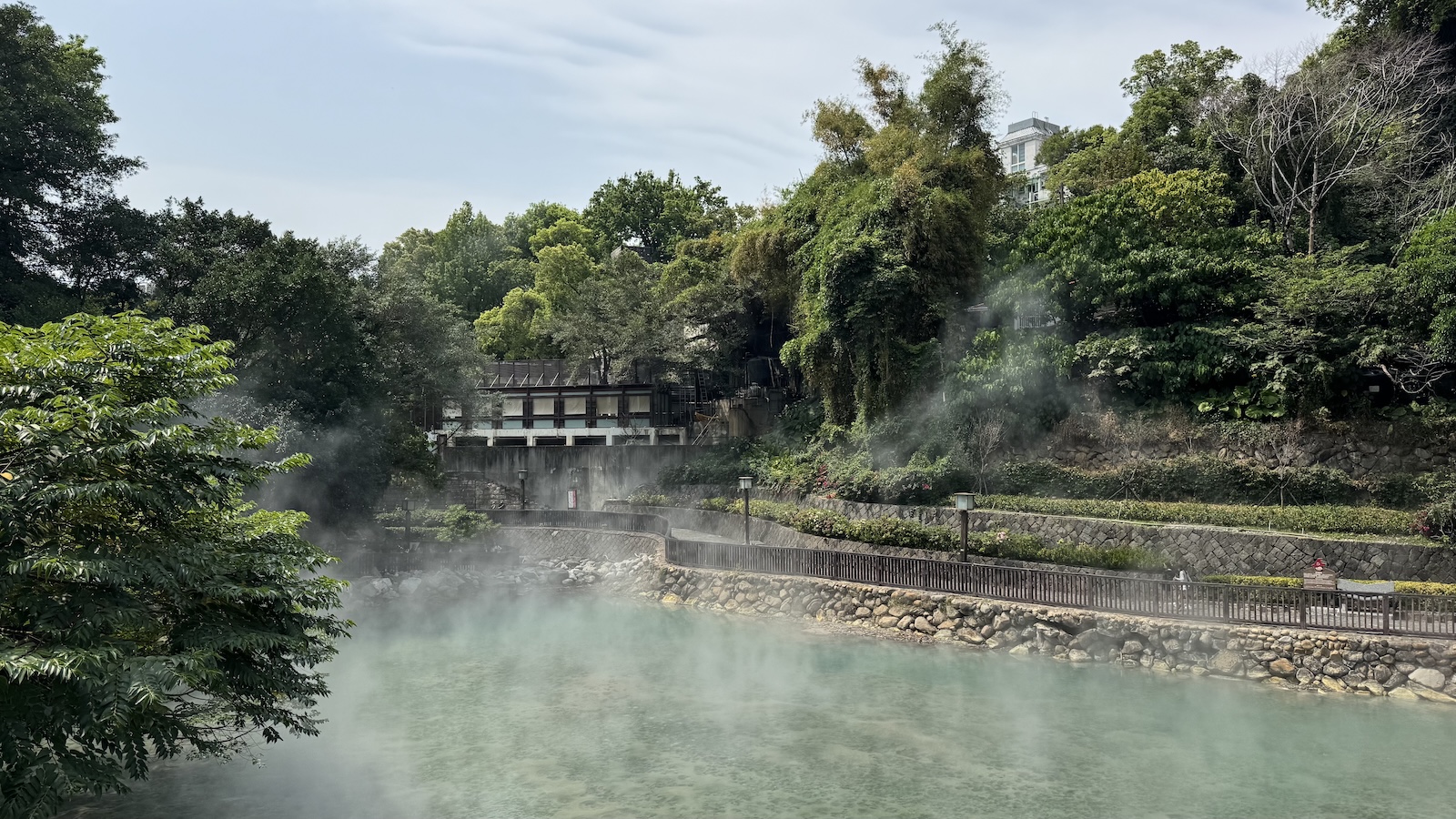 Steam rising from the thermal valley’s pale jade-green spring, framed by dense trees and a walkway of stone.