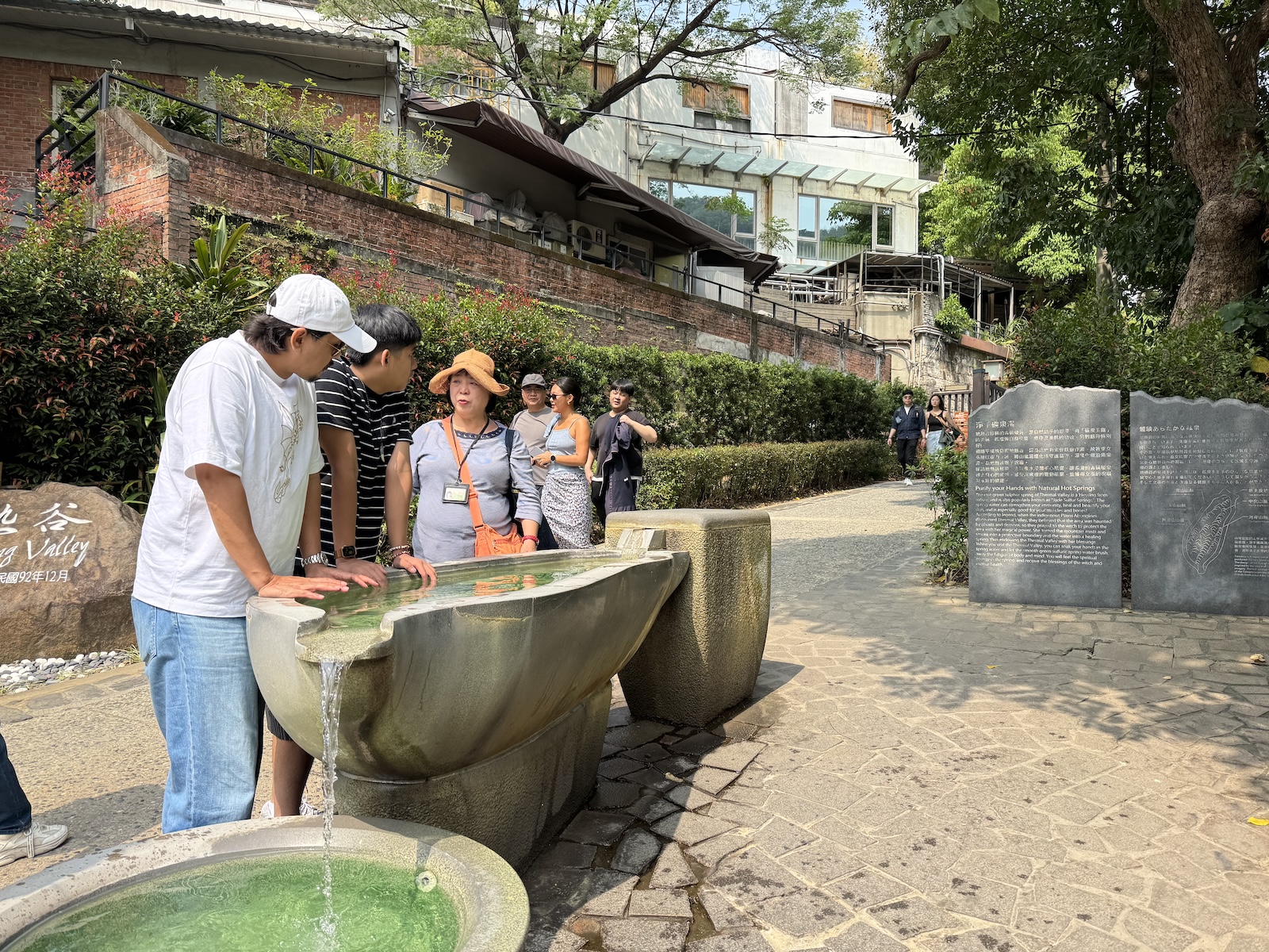 Visitors lean over a stone basin at Thermal Valley’s entrance to rinse their hands with spring water. Stone information panels describe the area.