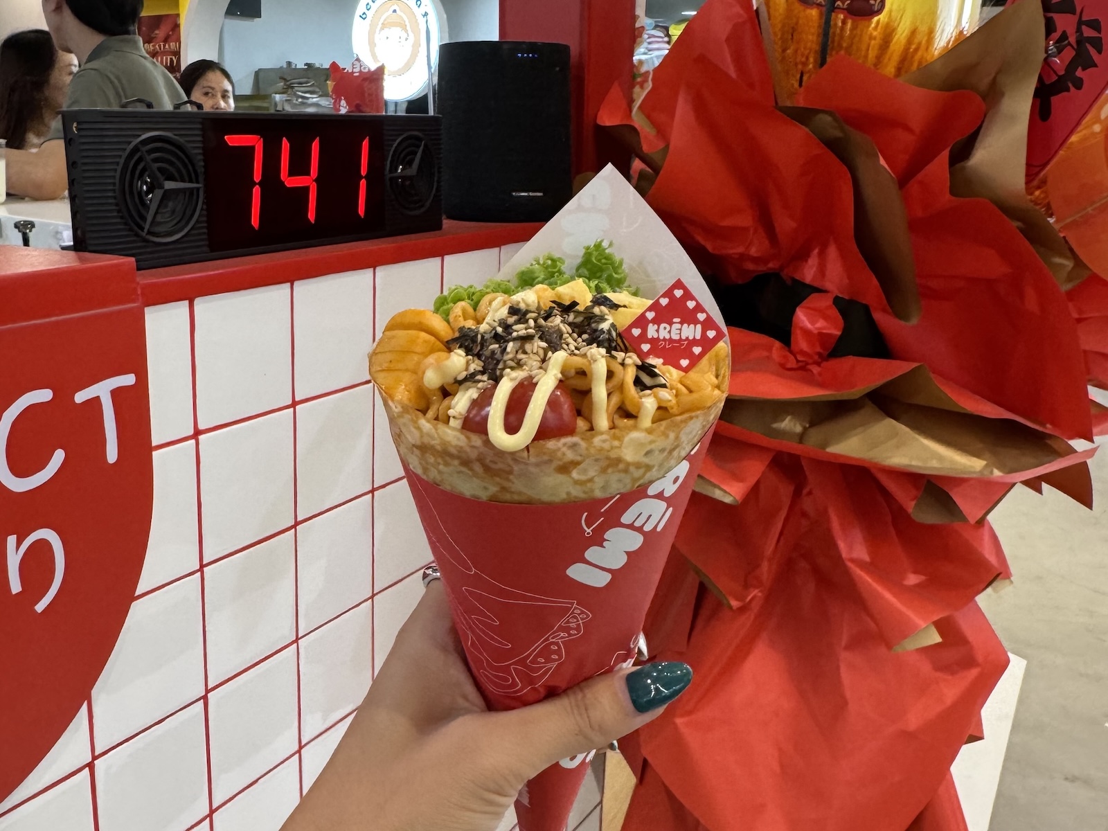 Hand holding a crepe cone filled with fried noodles and toppings, against a colourful diner-style backdrop.