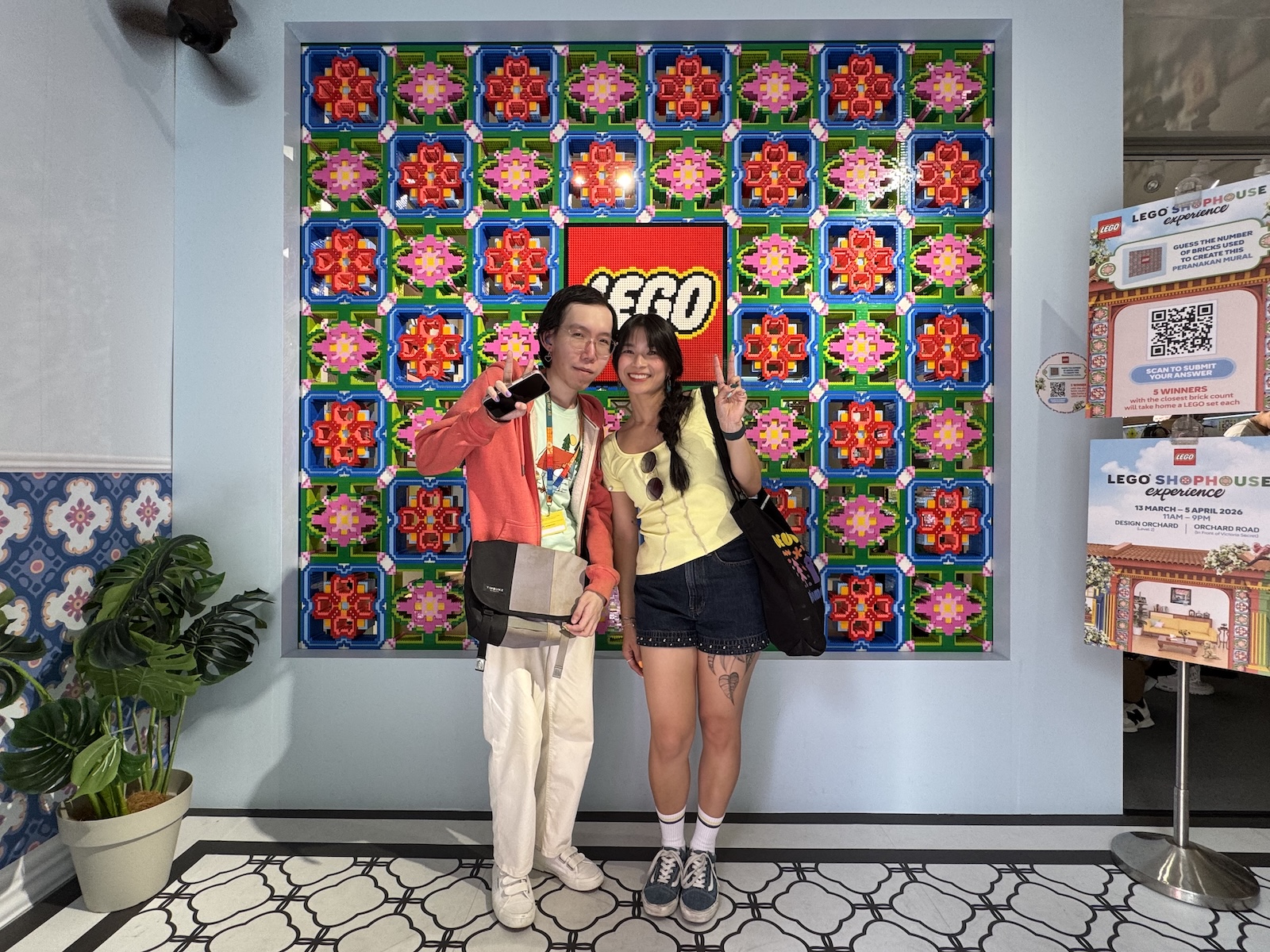 Two people pose before a colorful checkered patterned artwork, made of LEGO and resembling flowers. They wear a coral jacket with cream trousers and a yellow top with black shorts and a black bag.
