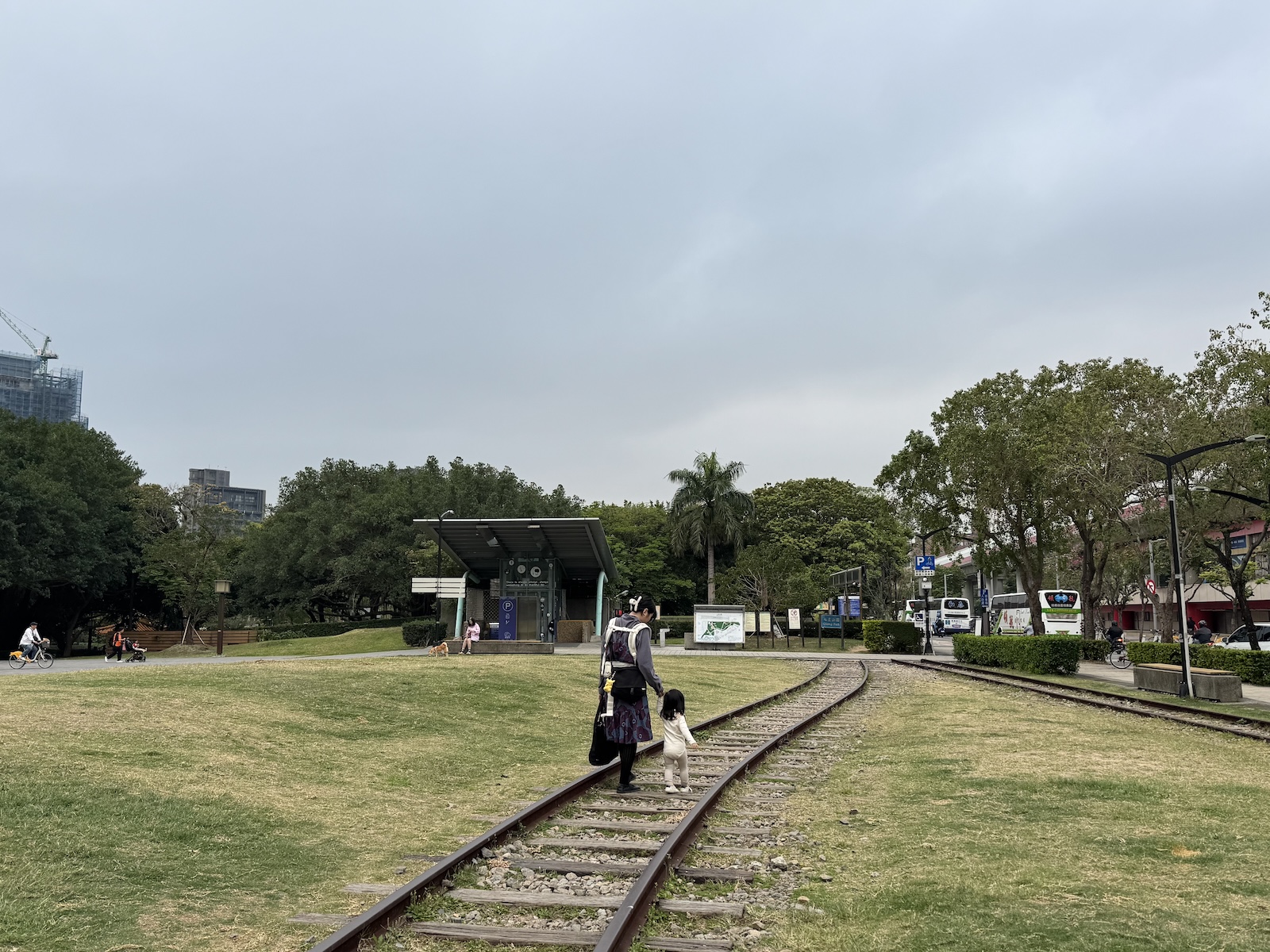 A mother and her child walk along the preserved railway tracks in Qixing Park near Xinbeitou Historic Station. The grassy park, trees, and station-side structures sit beneath an overcast sky