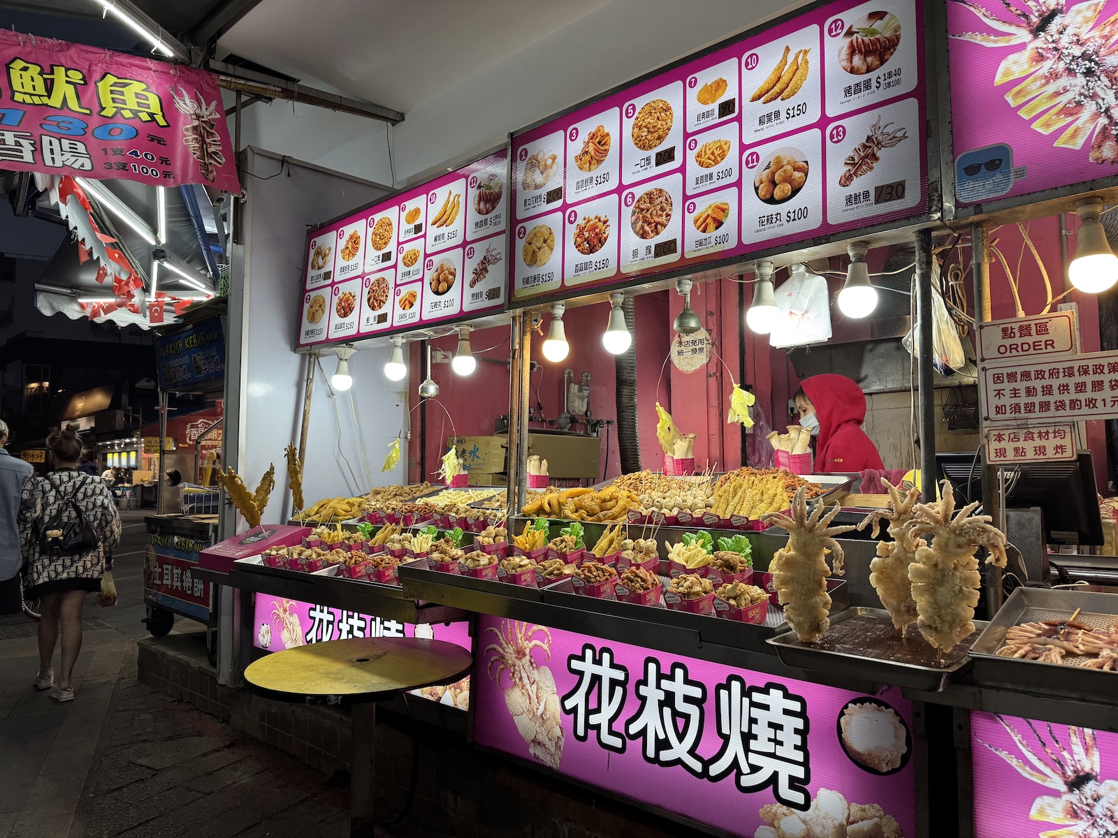A brightly lit stall displays skewered, golden-brown snacks beneath a photo menu, with shoppers passing by.