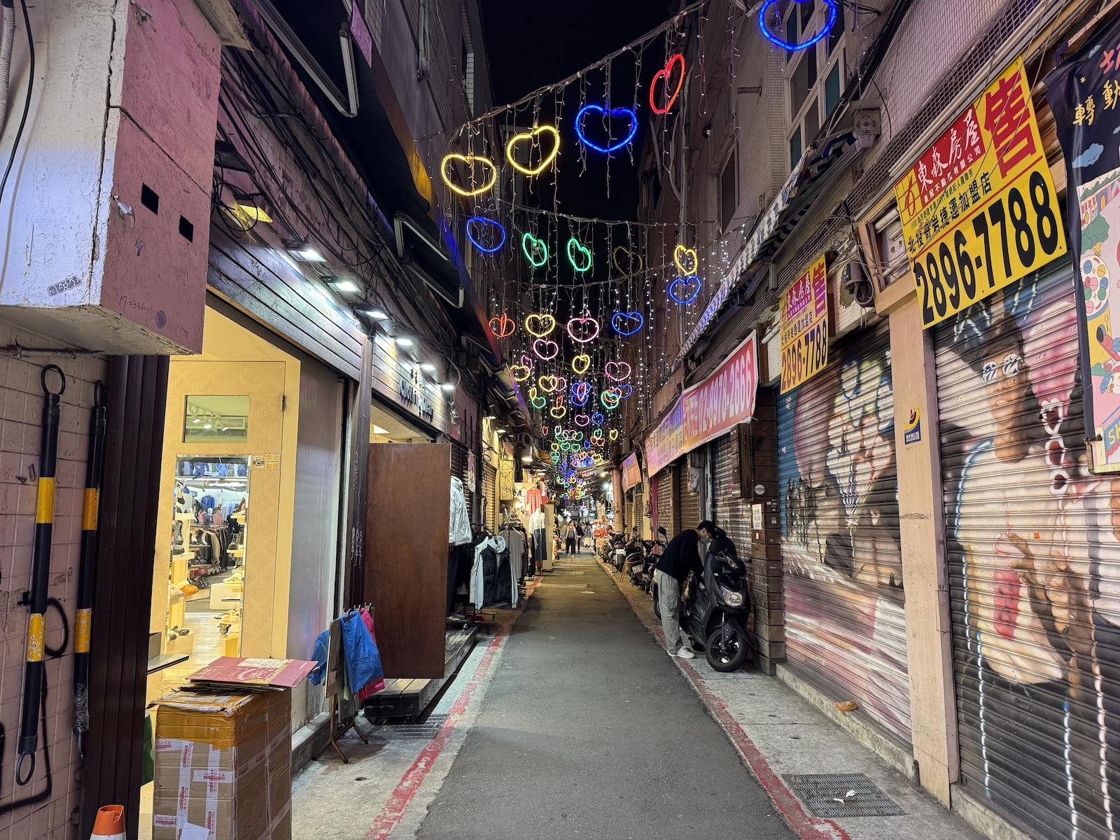 A narrow lane in Taipei’s Shilin Night Market recedes beneath colorful circular lights, with shopfronts, closed metal shutters, and a few people and scooters along the walkway. 