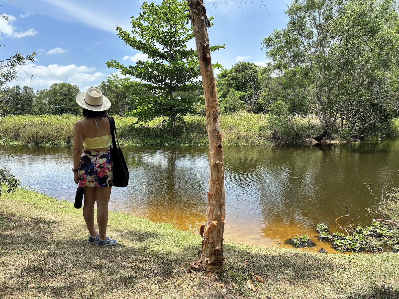 Me wearing a white sunhat, yellow crop top, floral shorts, and blue shoes and standing beside a pond.