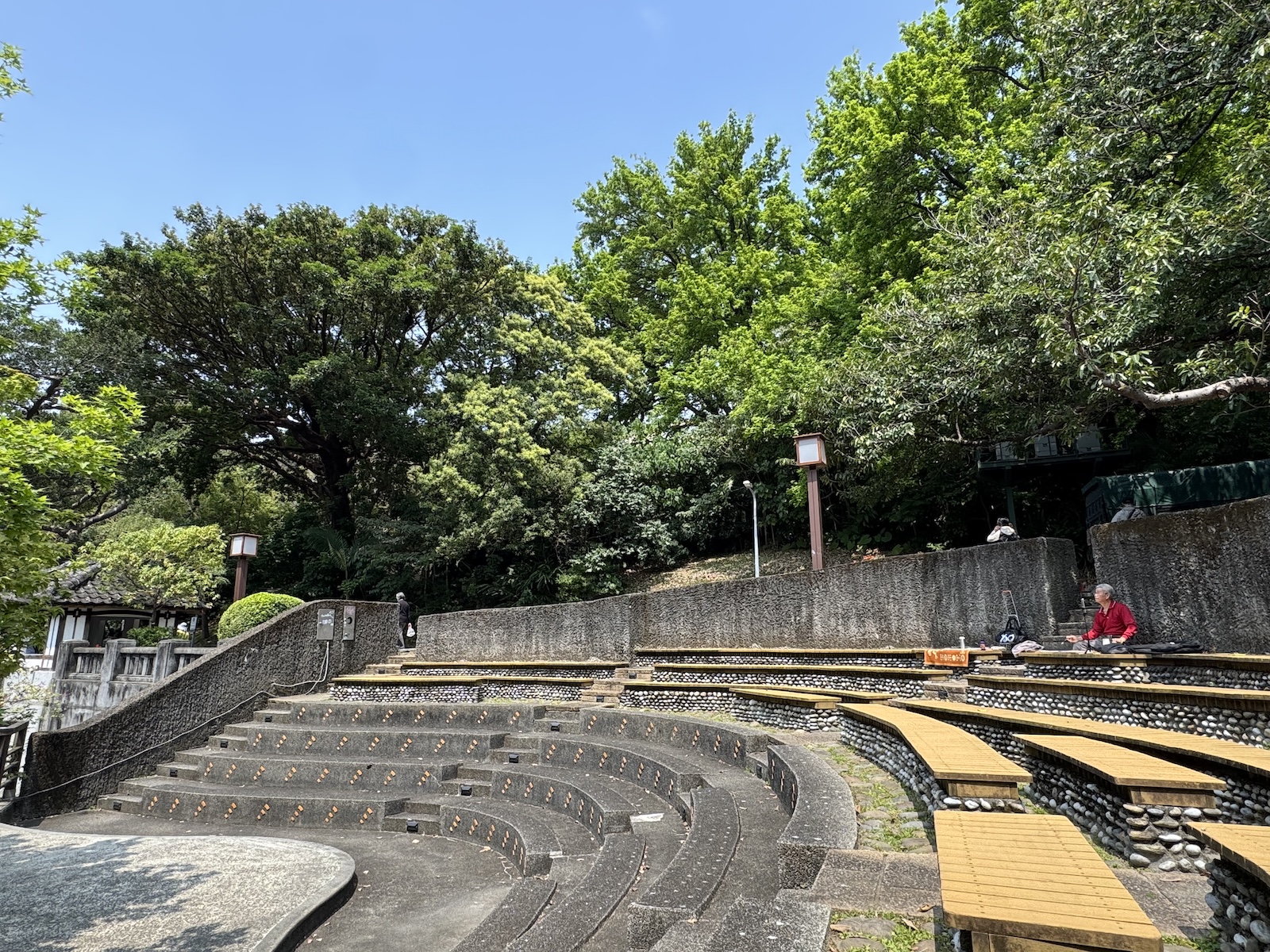 Curved rows of stone seating and small water outlets form an outdoor ampitheatre beneath the trees in Beitou Hot Spring Park. A visitor sits on the upper tier with a sketchbook.