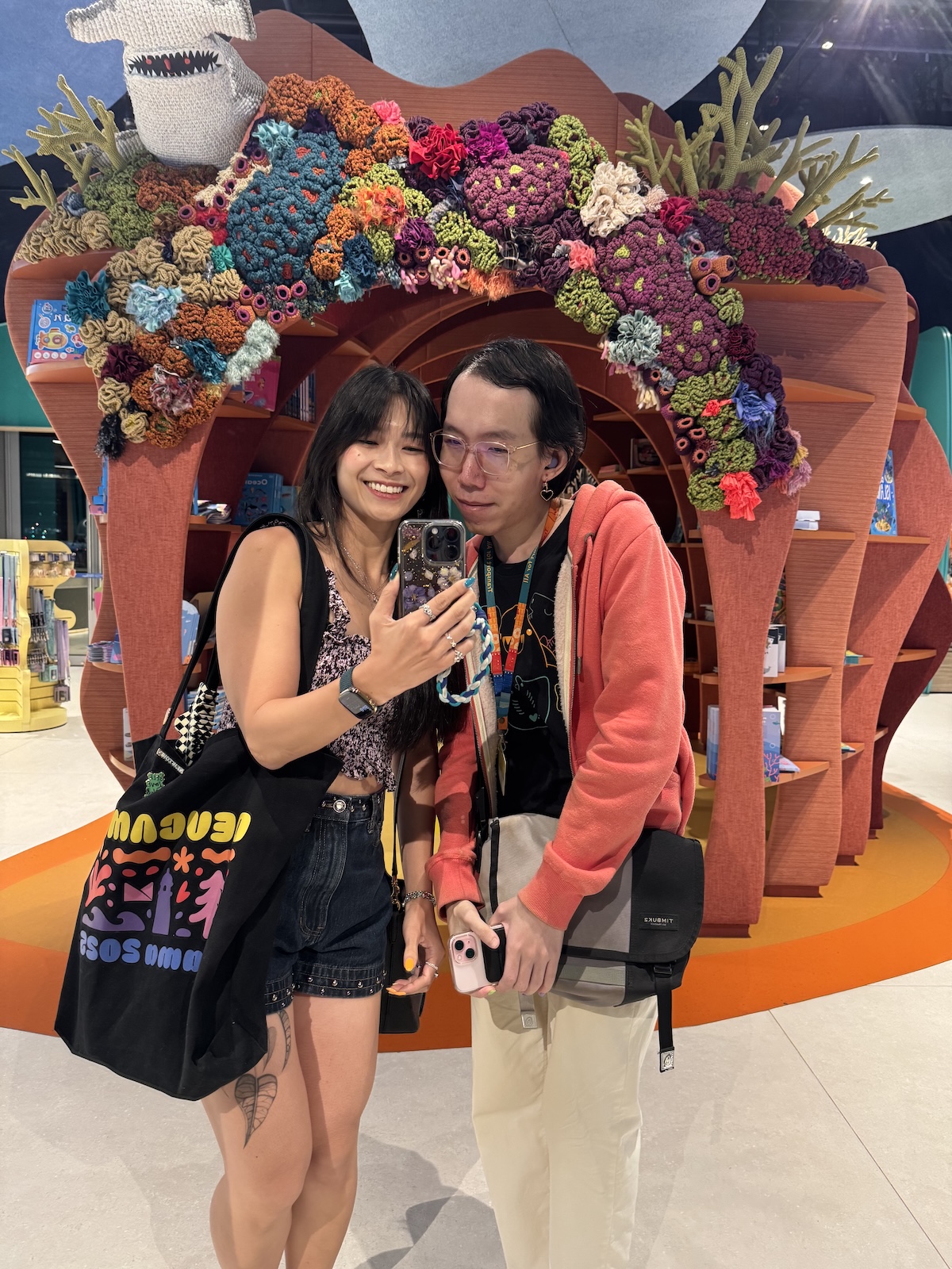 Me and Daniel posing for a selfie indoors in a mirror in front of a terracotta-coloured arch decorated with colourful flowers, and a hammerhead shark sculpture in the top left of the arch. I am wearing a black sleeveless top and denim shorts, and Daniel is wearing a coral jacket and beige trousers.