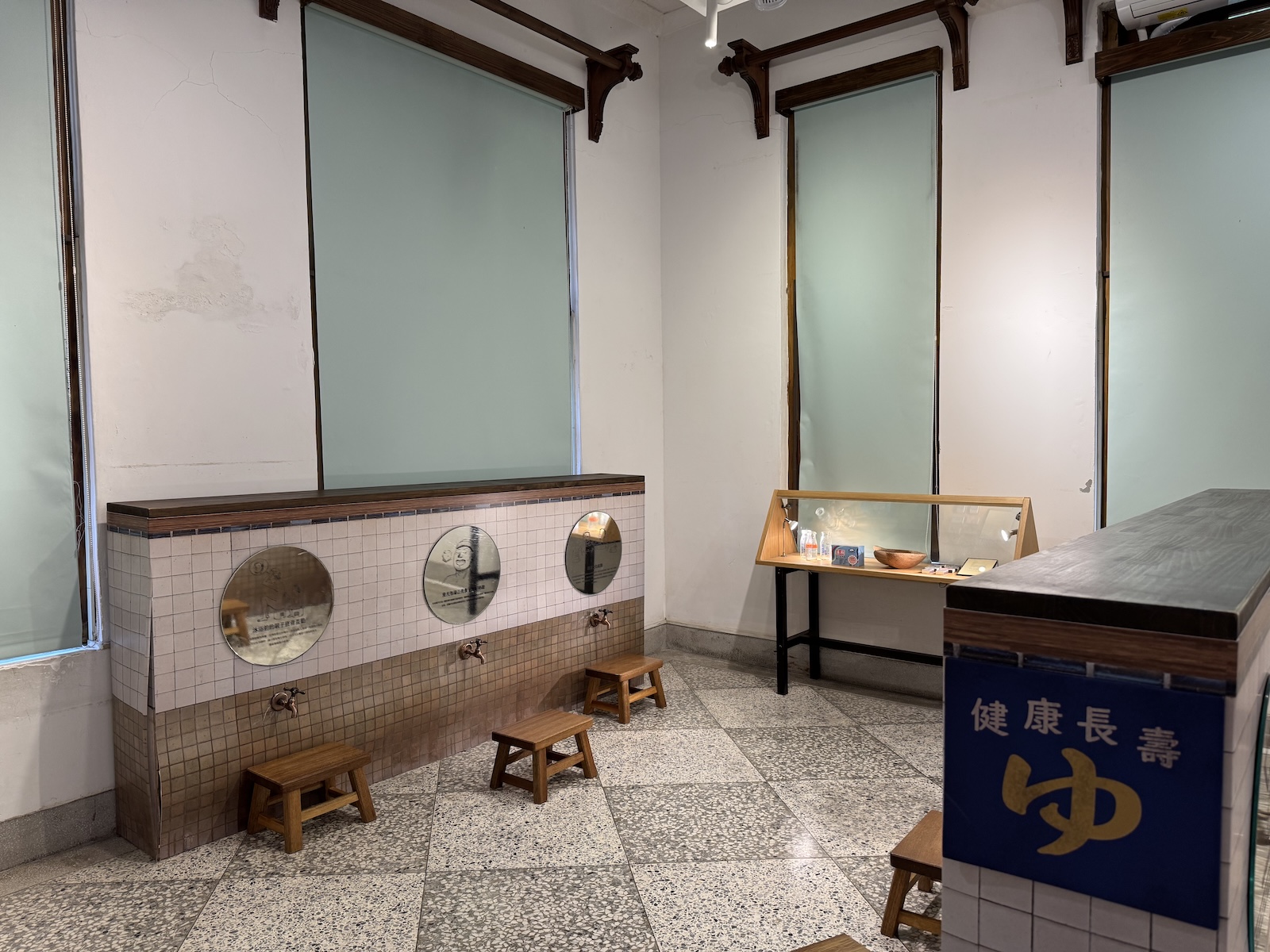 A tiled exhibit wall displaying old bathing fixtures and stools beside a glass case of artifacts, evoking the bathing rooms of the historic bathhouse.