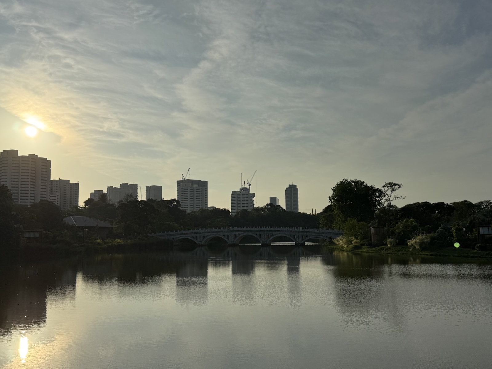 Sunset over a lake, with a low arched bridge, trees, and high-rise buildings reflected in the water.
