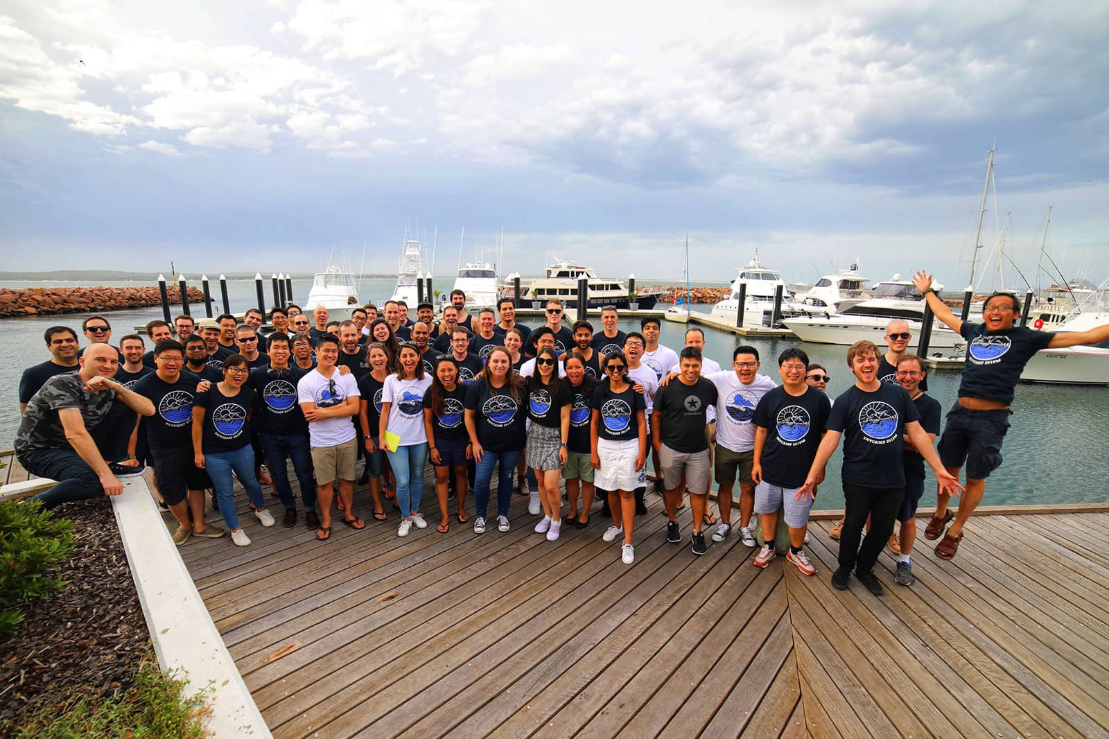 A group of people on a boardwalk with some small boats in the background. Many of the people are wearing the same t-shirt with a sun graphic in white and purple. The sky is a light blue colour with clouds