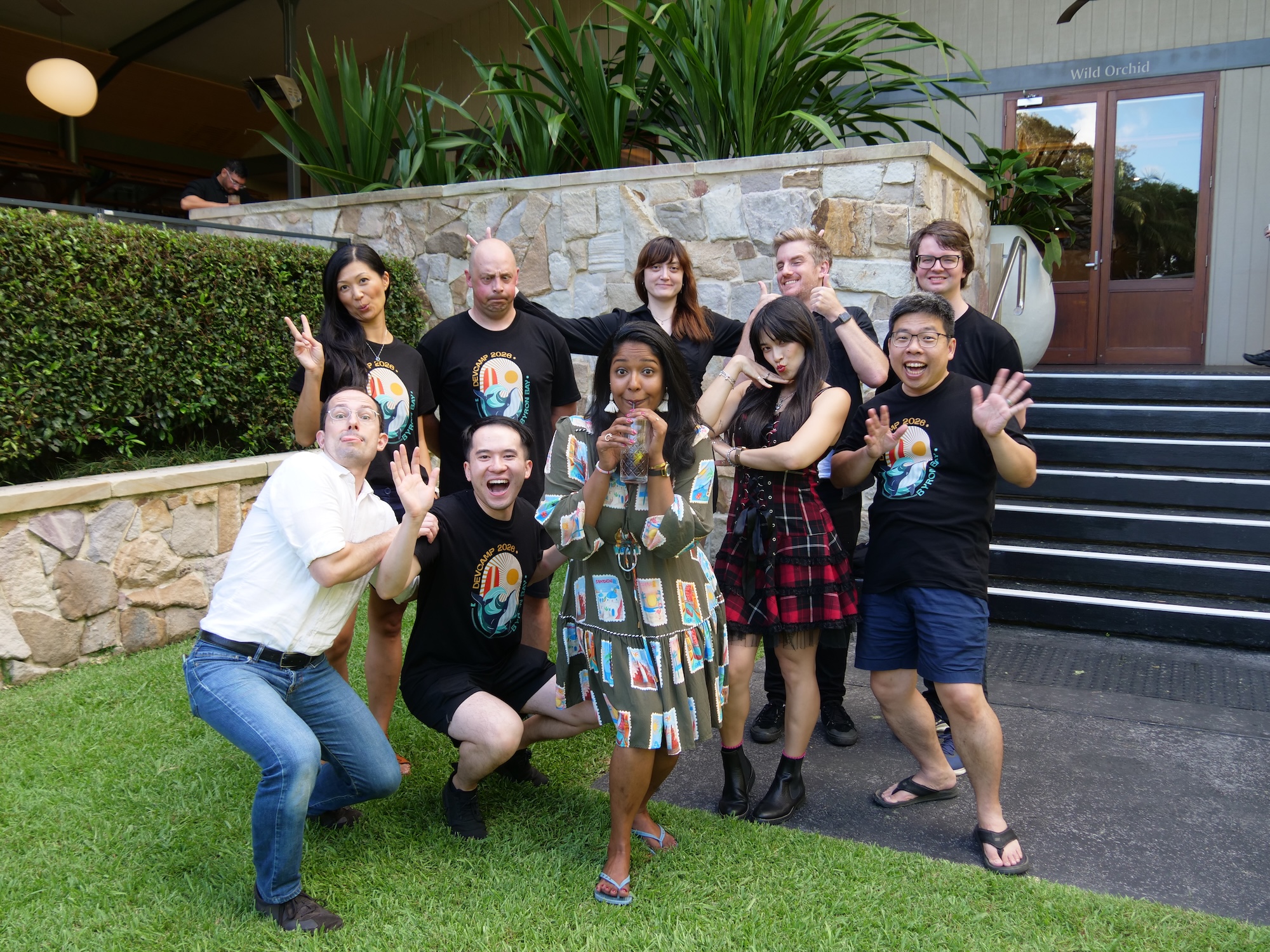 A group of ten people making silly faces at the camera and doing hand poses, standing on some concrete and well manicured bright green grass.