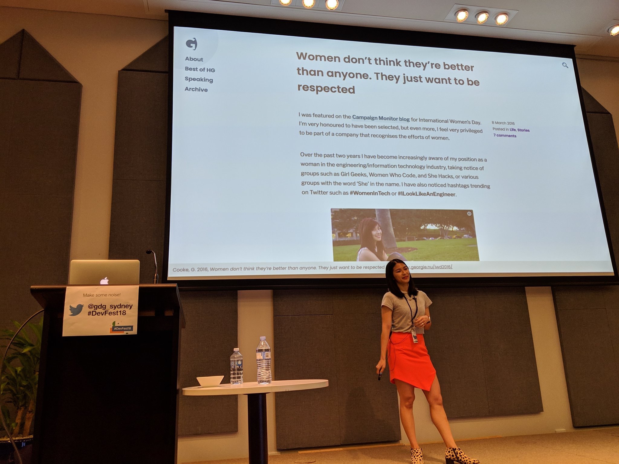 A person in a grey top and bright orange skirt and animal print boots, standing on a stage in front of a projector and next to a podium with a laptop. The screenshot on the projector has the title “Women don’t think they’re better than anyone. They just want to be respected”