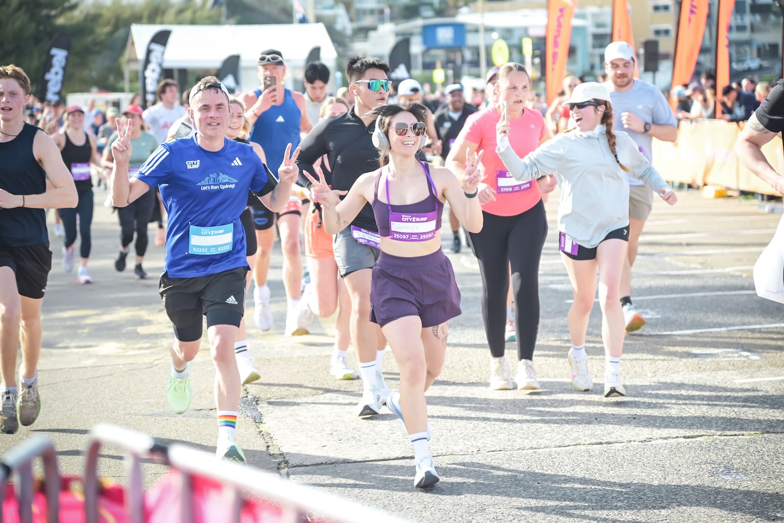 City2Surf participants run toward the finish area, including a smiling runner in a blue shirt and another person in sunglasses and purple athletic clothing waving beside them. Both are holding up the two-finger peace sign.