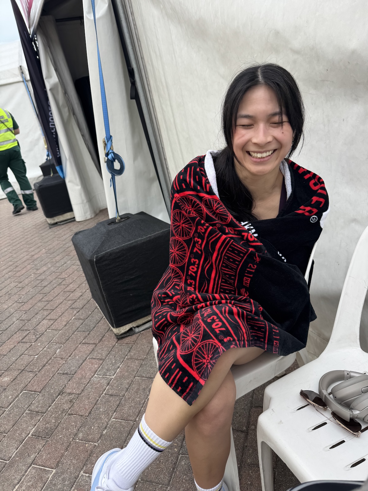 A person with dark hair smiling with their eyes slightly shut, with a black towel with a red pattern wrapped around them. They are wearing athletic socks and shoes. They are sitting on a plastic chair in front of a tent.