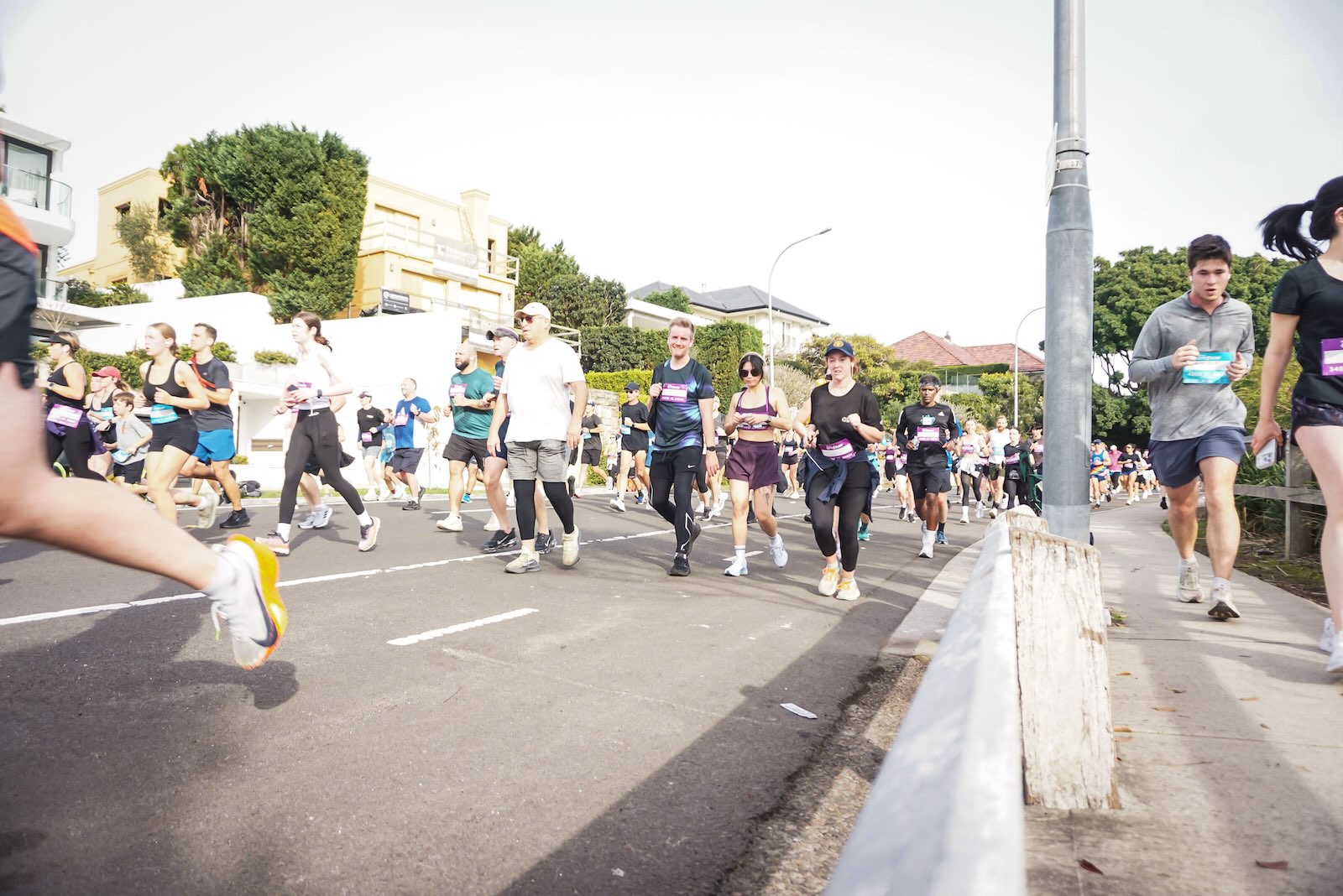 Runners and walkers take part in City2Surf on a broad residential street, with spectators along the footpath and a metal barrier in the foreground.