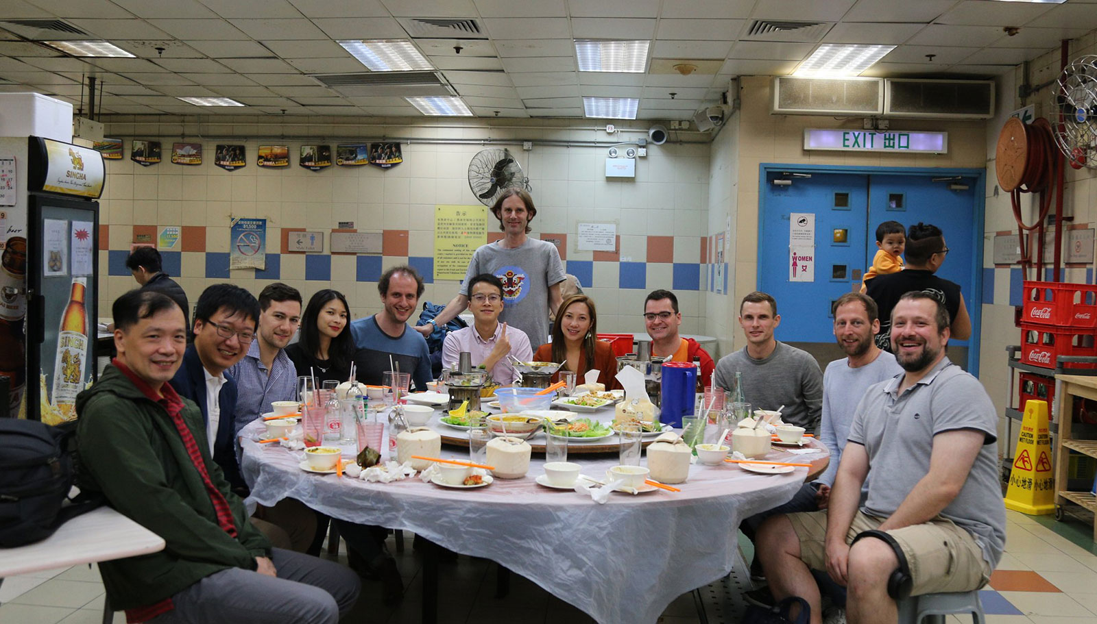 A group of people around a round table with a tablecloth, in a food court with a patterned tiled wall. There are lots of bowls and plates half-filled with food, and some fresh young coconut cut open with straws sitcking out of them