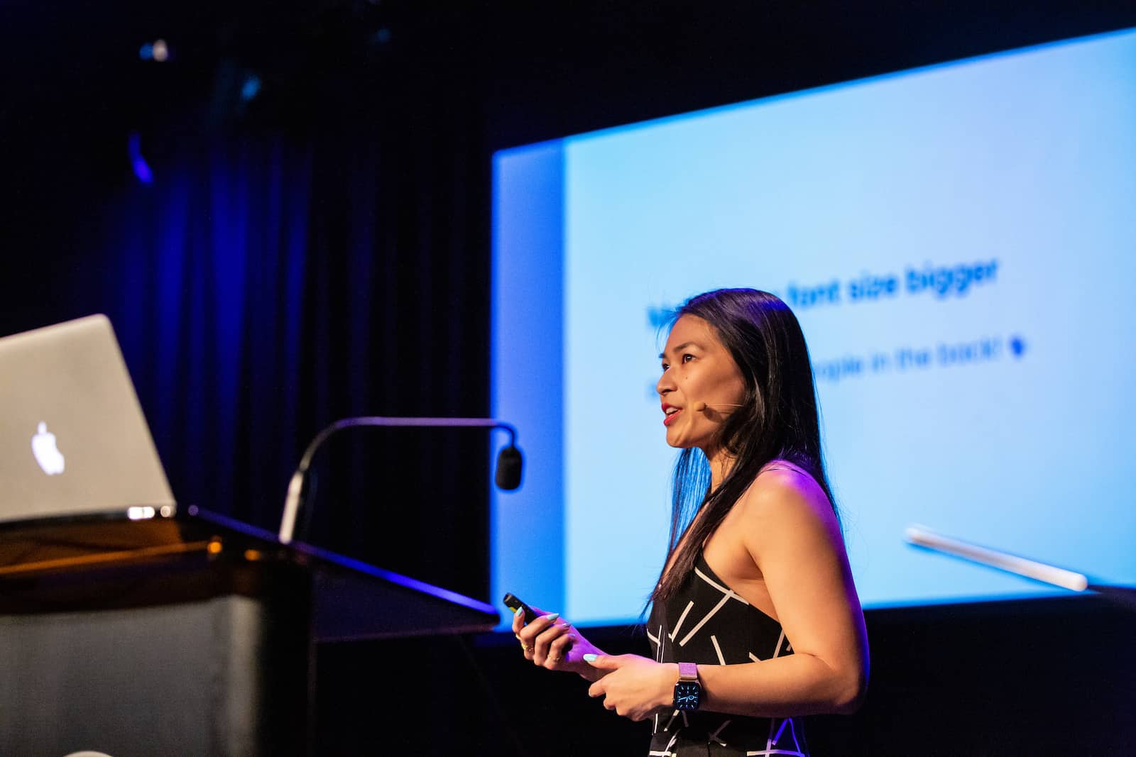 A person with long dark hair and a patterned sleeveless top, standing at a podium, with a headset microphone and holding a remote control. There is a projector in the background