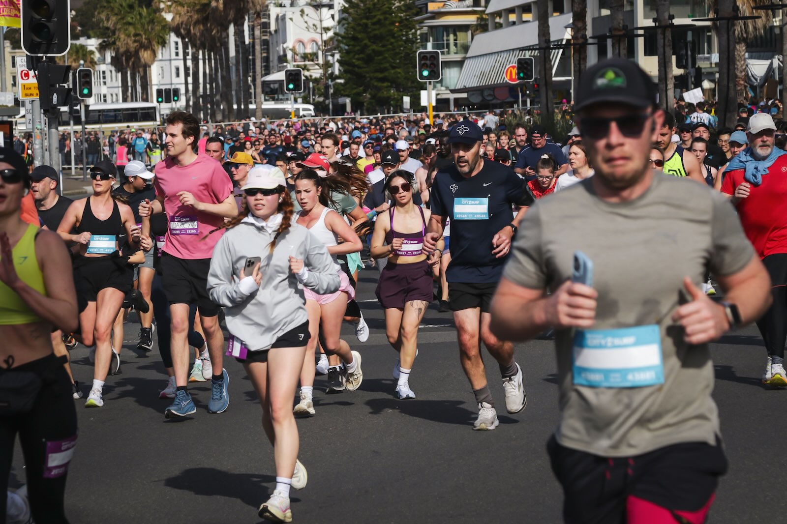 A large crowd of City2Surf runners fills a city street lined with palm trees, traffic lights, and spectators