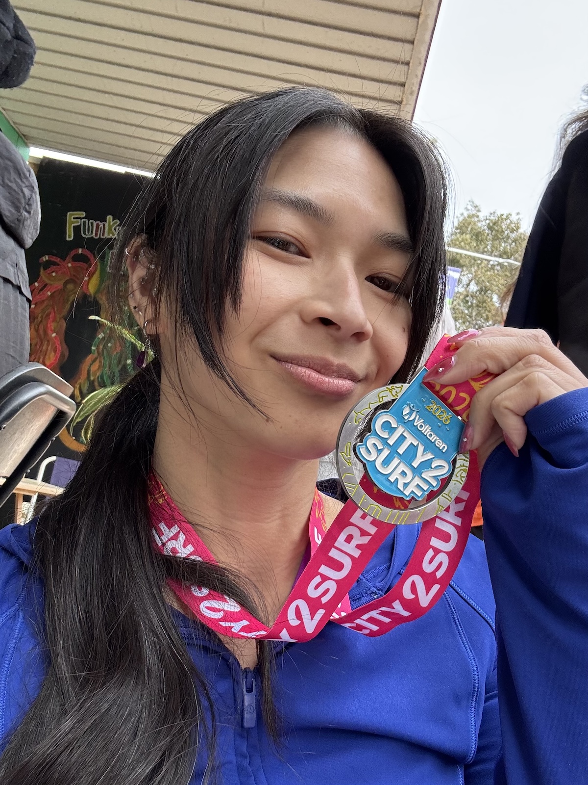 A person with long dark hair, wearing a blue top and a medal around their neck with a pink lanyard. The lanyard and the medal read City2Surf