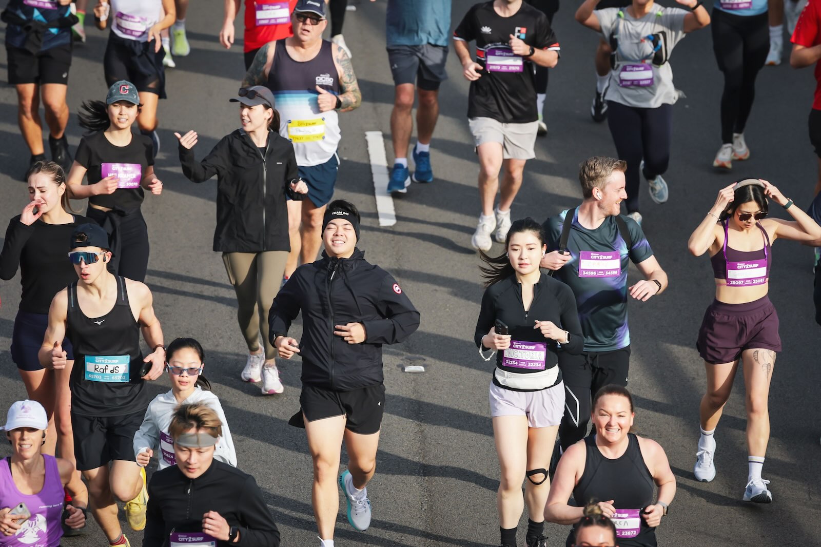 An overhead view shows a diverse group of City2Surf runners moving along the road, wearing event bibs and varied athletic clothing.