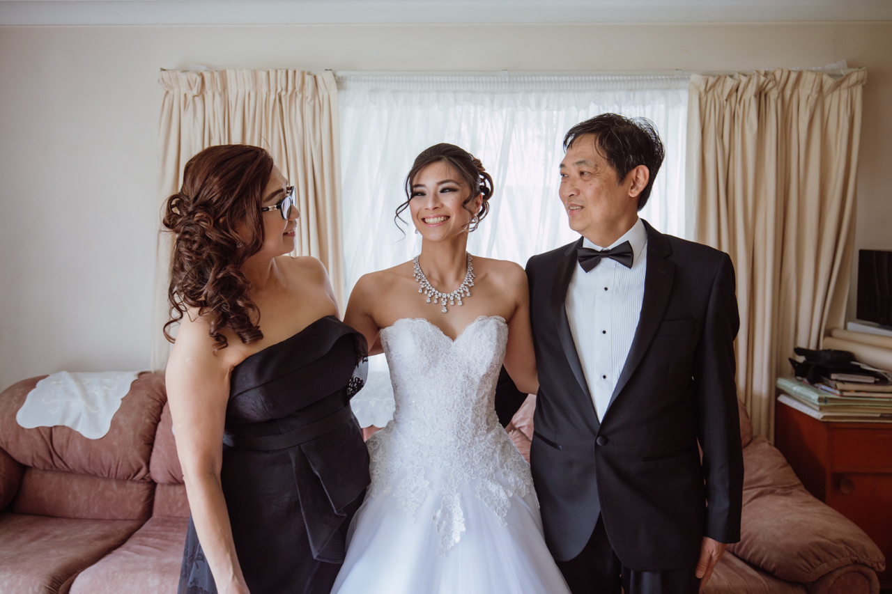 A woman in a white strapless wedding dress standing between her mum and dad, dressed in a black dress and a black suit, respectively. They are looking at each other