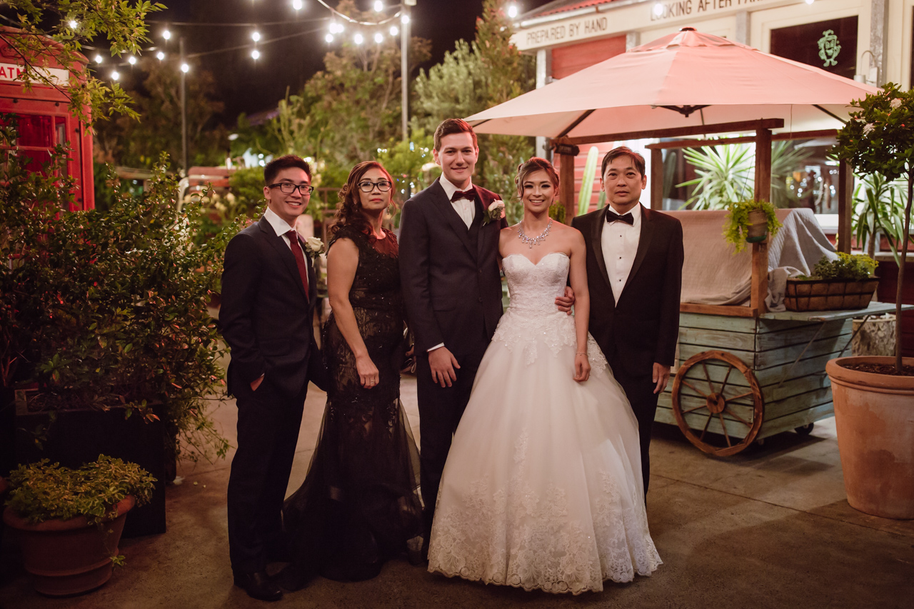 A married couple, the groom in a navy suit and the bride in a white ballgown, along with the bride’s family – mum, dad, and brother. It’s evening and they are outside in a garden-like setting